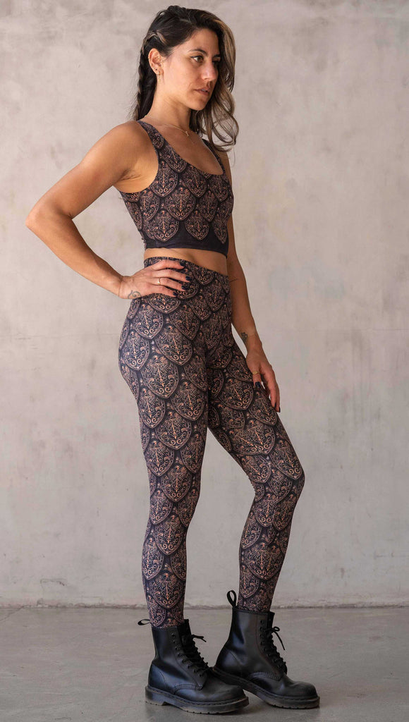 Full body side view of model wearing WERKSHOP Dragon Rider leggings in Gold. The artwork features an intricate battle shield designed to look like dragon scales. This color way is all shades of gold and bronze.