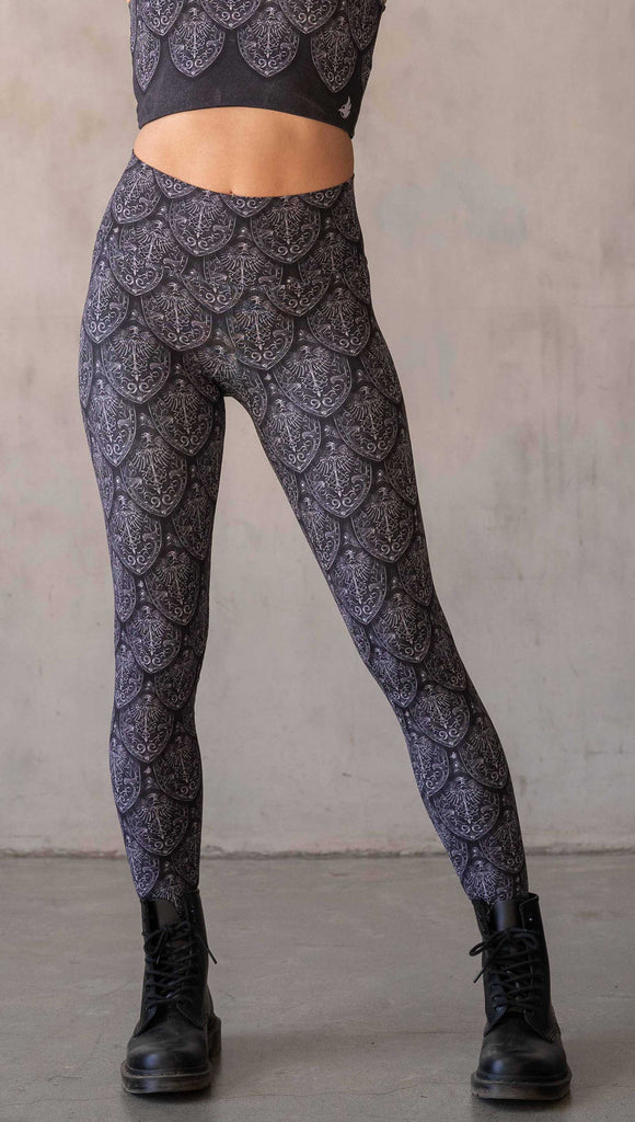 Front view of model wearing WERKSHOP Dragon Rider leggings in Silver. The artwork features an intricate battle shield designed to look like dragon scales. This color way is all shades of black and gray.