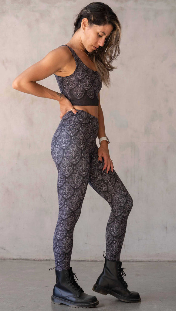 Full body view of model wearing WERKSHOP Dragon Rider leggings in Silver. The artwork features an intricate battle shield designed to look like dragon scales. This color way is all shades of black and gray.