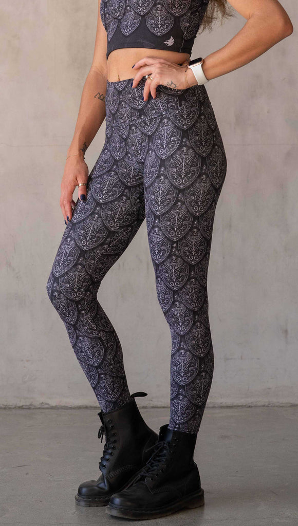 Side view of model wearing WERKSHOP Dragon Rider leggings in Silver. The artwork features an intricate battle shield designed to look like dragon scales. This color way is all shades of black and gray.