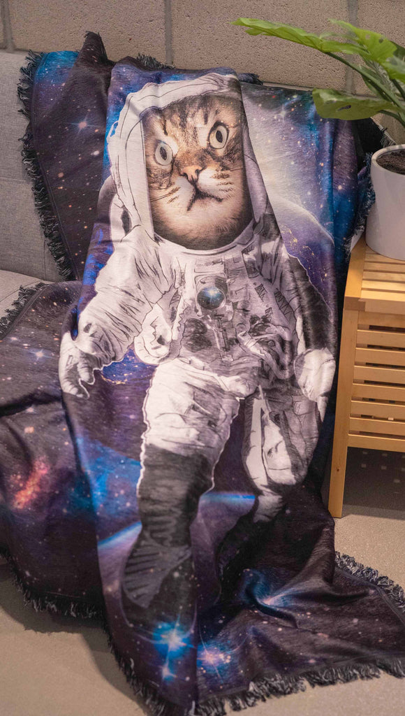 Decorative Chenille Tapestry printed with limited edition artwork by our Female Founder, Chriztina Marie. The artwork features an adorable domestic house cat wearing an astronaut uniform, floating in outer space with a nebula behind him. This photo shows the tapestry draped over the edge of a couch.