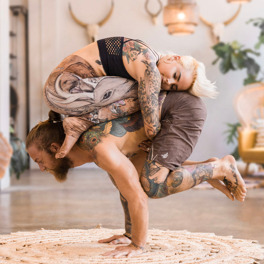 A couple doing "koalasana" acro yoga pose with a girl balancing on a boys back while he is in crow pose. They are both wearing WERKSHOP head-to-toe