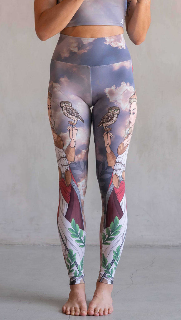 Front view of model wearing WERKSHOP Athena leggings. The leggings are printed with original artwork by Chriztina Marie and features Athena, the goddess of war standing on a cliff’s edge. She is holding a spear with one hand and her owl with the other. She is wearing a greek goddess dress/warrior hat with colors of cream, gold and red. Behind her is a beautiful cloudy sky with rays of light shining down onto her. The model is also wearing a matching top.