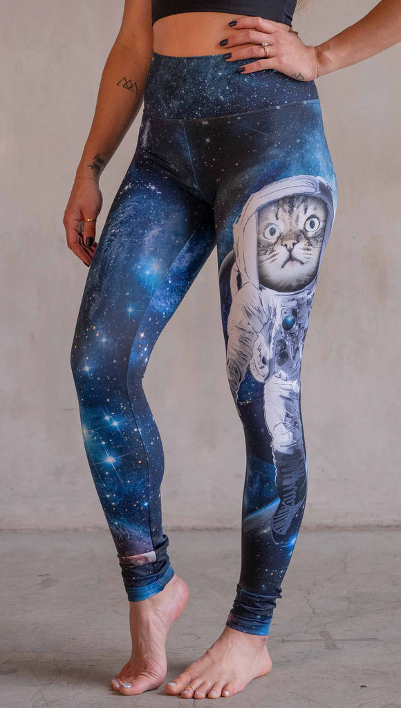 Model wearing WERKSHOP Catstronaut Leggings. The leggings are printed with a cat in an astronaut suit on the wearer's left leg. The galaxy background is a deep cobalt blue with nebula swirls and shooting stars.