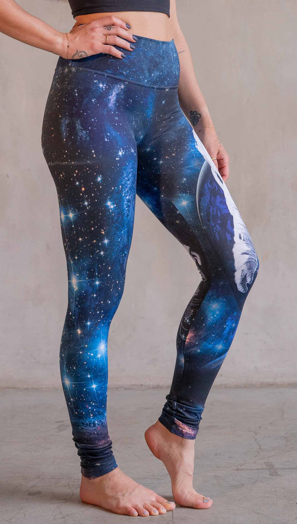 Model wearing WERKSHOP Catstronaut Leggings. The leggings are printed with a cat in an astronaut suit on the wearer's left leg. The galaxy background is a deep cobalt blue with nebula swirls and shooting stars.