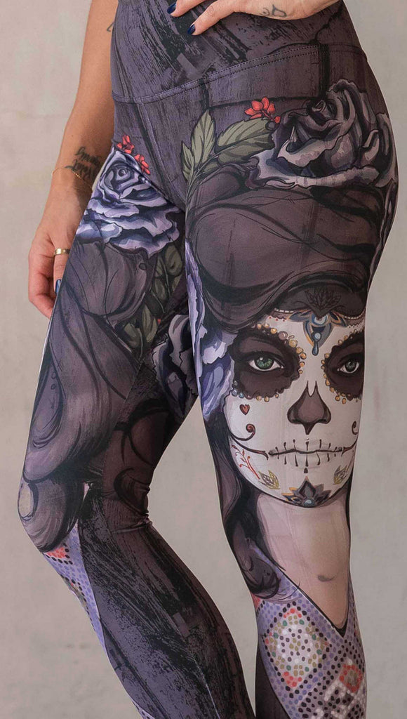 Girl wearing WERKSHOP "Dark Sugar" Athleisure leggings. The artwork on the leggings features a girl wearing dia de los muertos makeup in tones of blue and gray. she is wearing a crown of roses and a beaded necklace over a distressed warm gray woodgrain background.