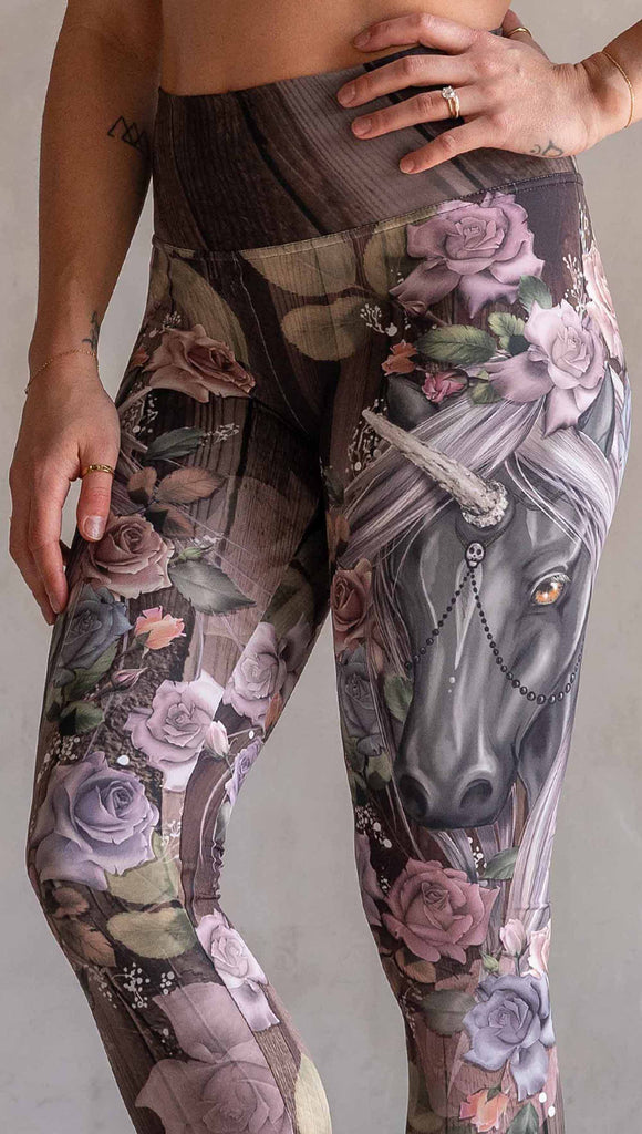 Zoomed in view of model wearing WERKSHOP Dark Unicorn Athleisure Leggings. The leggings feature original artwork of a unicorn surrounded by a wreath of vintage coloured roses. The unicorn has a beaded chain with a skull dangling from it's horn. The artwork is over a warm woodgrain background.