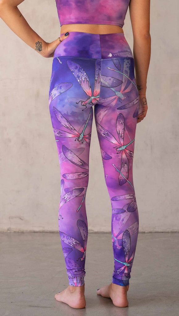 Back view of model wearing WERKSHOP Dragonflies Athleisure leggings. The artwork on the leggings features hand drawn dragonflies with pops of teal and pink over a purple and pink watercolour background.