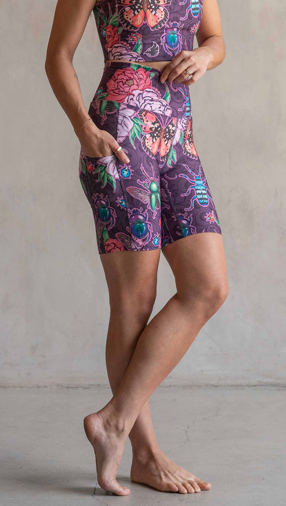 Side view of model wearing WERKSHOP Enchanted Garden EnviSoft Bicycle length shorts with pockets. The fabric is printed with original artwork by our Female Founder, Chriztina Marie. The artwork printed not the fabric features Butterflies, Beetles and Peonies over a warm fuchsia with bright bold pops of color on each beetle and Butterfly.
