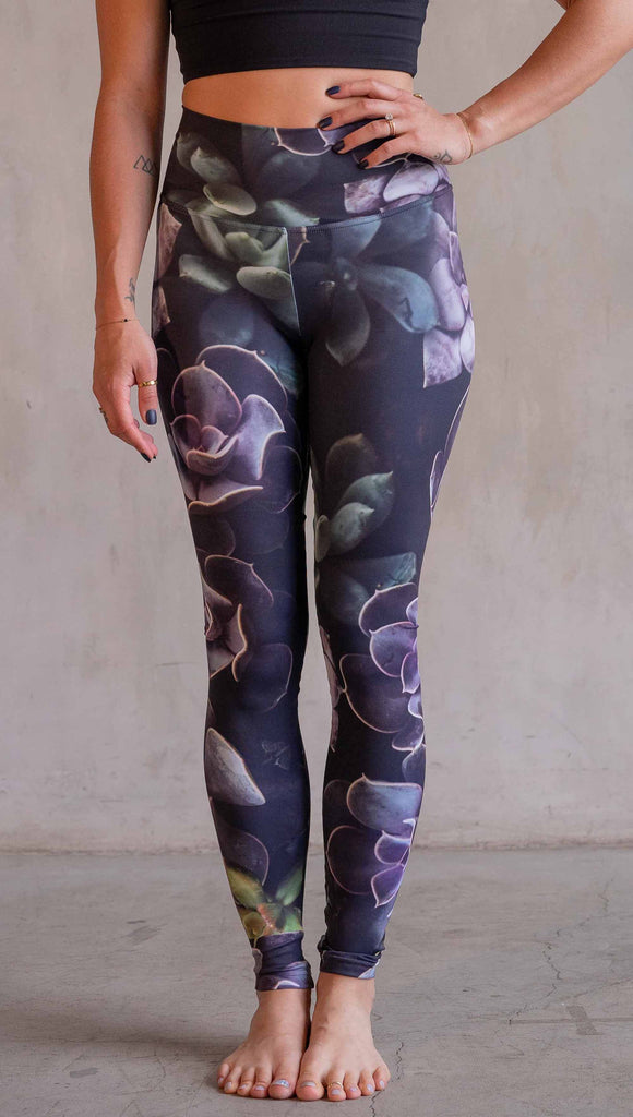 Model wearing WERKSHOP Midnight Garden Athleisure Leggings. The artwork on the leggings is printed with succulents in dark romantic colors of purple with small pops of teal