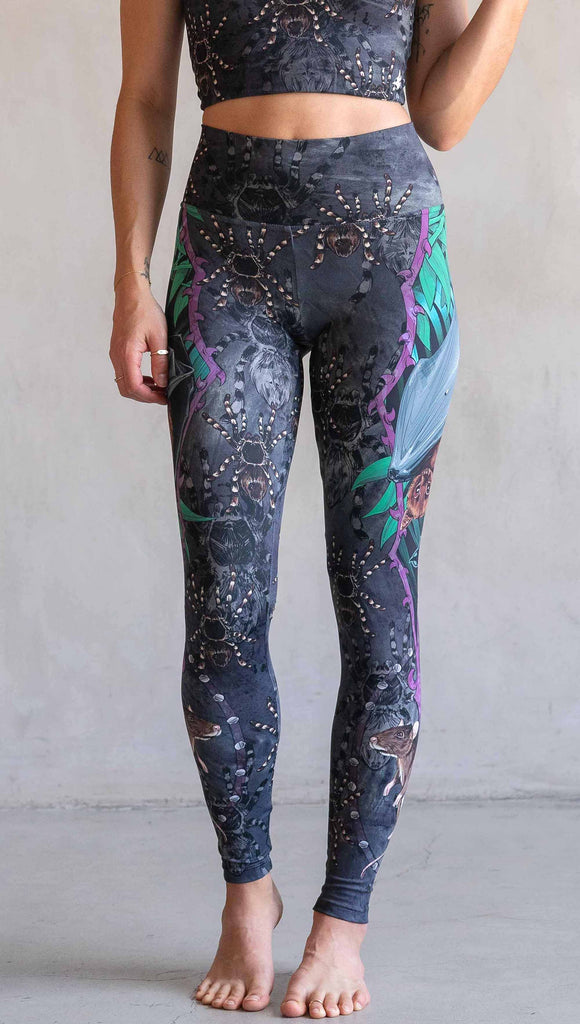 Model wearing WERKSHOP Spooky Season Set. The leggings feature an adorable fruit  bat dangling upside down inside a tropical scene with a purple wreath of thorns. under the bat, there is a rat facing forward. The background is a distressed dark gray brushstroke texture with scattered tarantulas.