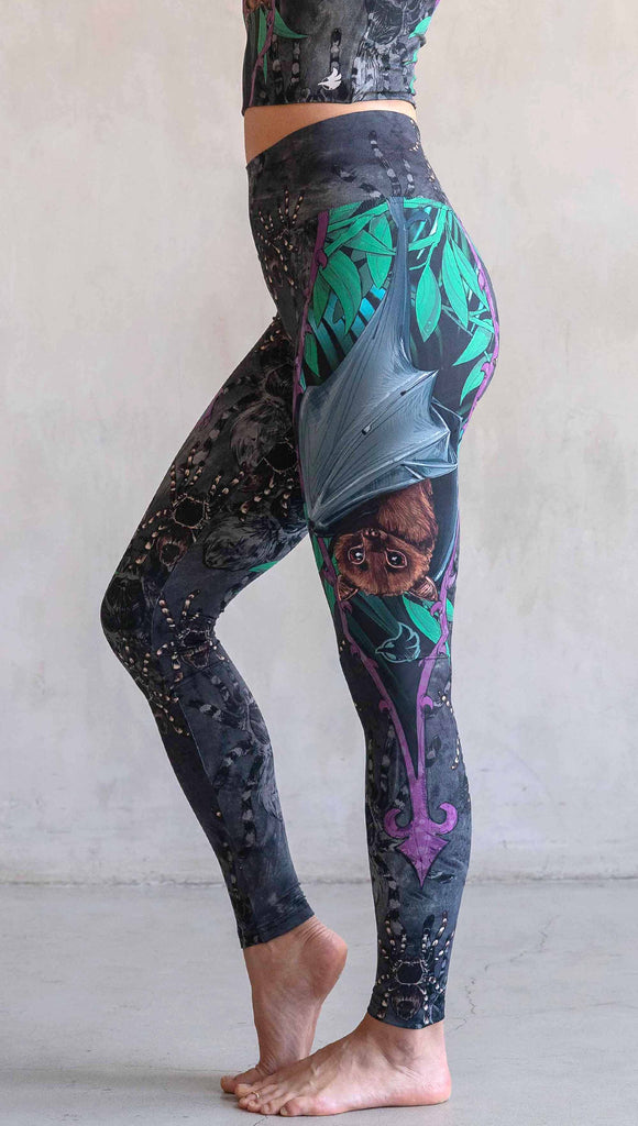 Model wearing WERKSHOP Spooky Season Set. The leggings feature an adorable fruit bat dangling upside down inside a tropical scene with a purple wreath of thorns. The background is a distressed dark gray brushstroke texture. (This version of the artwork has no rats)
