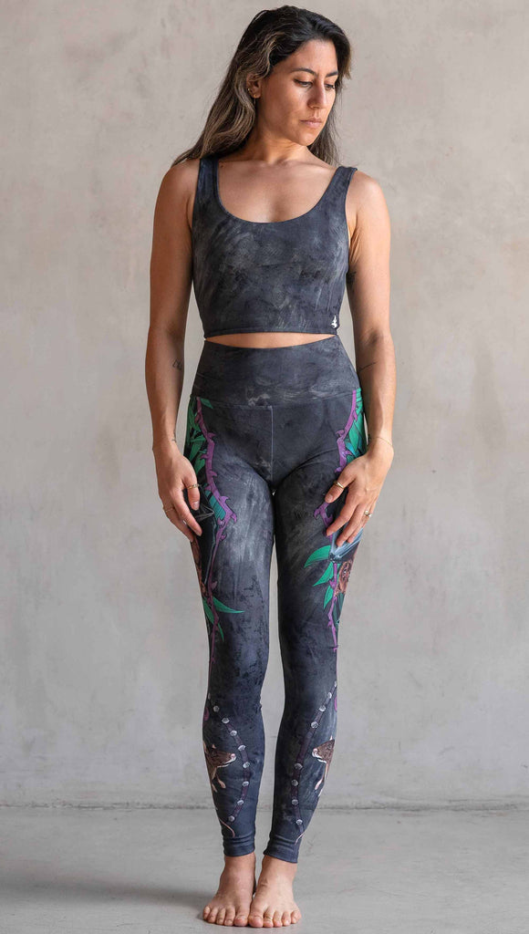 Model wearing WERKSHOP Spooky Season Set. The leggings feature an adorable fruit bat dangling upside down inside a tropical scene with a purple wreath of thorns. under the bat, there is a rat facing forward. The background is a distressed dark gray brushstroke texture. (This version of the artwork has no spiders)