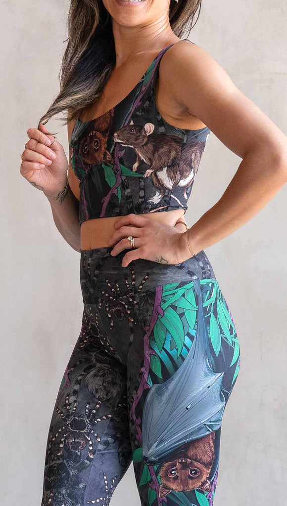 Zoomed in photo of model wearing WERKSHOP Spooky Season Set. The leggings feature an adorable fruit  bat dangling upside down inside a tropical scene with a purple wreath of thorns. under the bat, there is a rat facing forward. The background is a distressed dark gray brushstroke texture with scattered tarantulas.