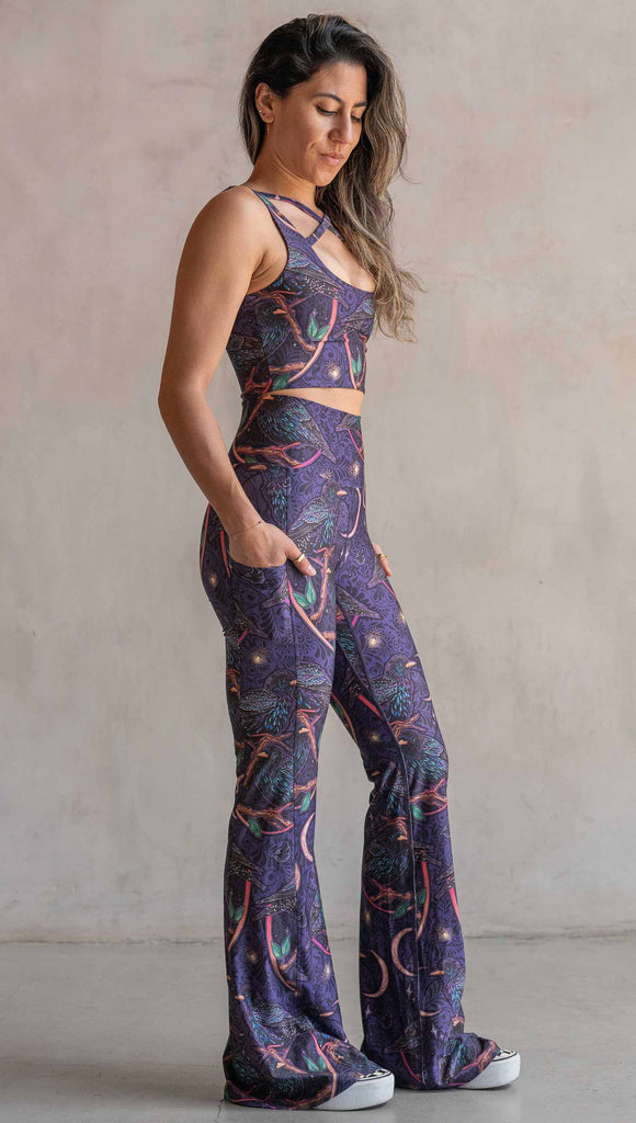Full body side view of model wearing WERKSHOP Starlings EnviSoft bell bottom pants with pockets. The fabric is printed with original artwork by our Female Founder, Chriztina Marie. Featuring European Starlings perched on a branch near a crescent moon and fireflies. The colors are warm purples with pops of pink, gold and green.