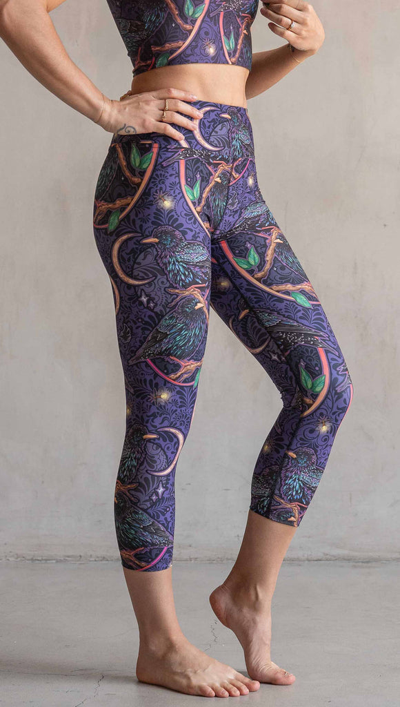 Side view of model wearing WERKSHOP Starlings Triathlon Capri length leggings. The fabric is printed with original artwork by our Female Founder, Chriztina Marie. Featuring European Starlings perched on a branch near a crescent moon and fireflies. The colors are warm purples with pops of pink, gold and green.