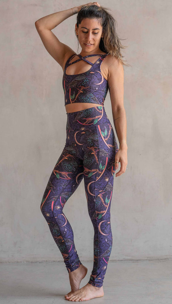 Full body view of model wearing WERKSHOP Starlings EnviSoft leggings with pockets. The fabric is printed with original artwork by our Female Founder, Chriztina Marie. Featuring European Starlings perched on a branch near a crescent moon and fireflies. The colors are warm purples with pops of pink, gold and green.