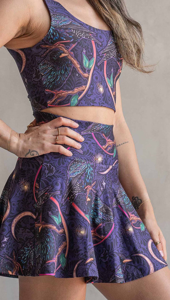 Zoomed in view of model wearing WERKSHOP Starlings EnviSoft Active Skirt with hidden shorts and pockets. The fabric is printed with original artwork by our Female Founder, Chriztina Marie. Featuring European Starlings perched on a branch near a crescent moon and fireflies. The colors are warm purples with pops of pink, gold and green.