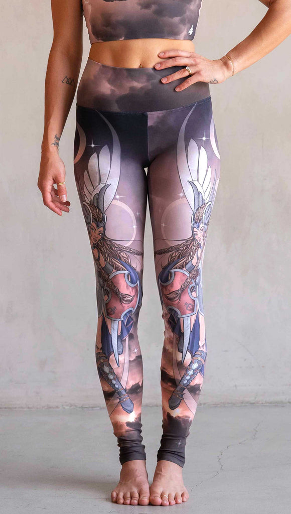 Waist-down front view of model wearing WERKSHOP Valkyrie Athleisure Leggings. featuring original artwork by Chriztina Marie. featuring a Valkyrie warrior from Norse Mythology flying through a dark sky holding a shield and swords. Her hair is in braids and she is wearing armor and a viking helmet. The colors are warm and the background also features a crescent moon and stars.