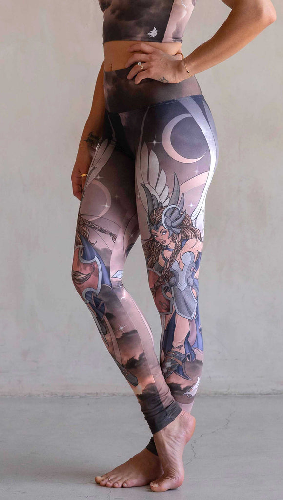 Side view of model wearing WERKSHOP Valkyrie Athleisure Leggings.  featuring original artwork by Chriztina Marie. featuring a Valkyrie warrior from Norse Mythology flying through a dark sky holding a shield and swords. Her hair is in braids and she is wearing armor and a viking helmet. The colors are warm and the background also features a crescent moon and stars.