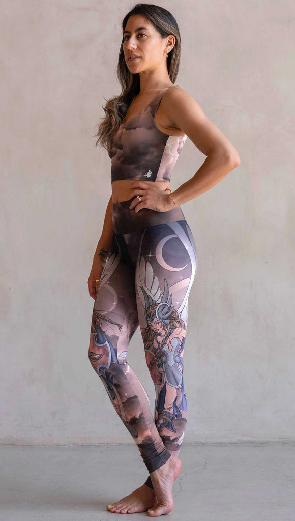 Full body side view of model wearing WERKSHOP Valkyrie Athleisure Leggings. featuring original artwork by Chriztina Marie. featuring a Valkyrie warrior from Norse Mythology flying through a dark sky holding a shield and swords. Her hair is in braids and she is wearing armor and a viking helmet. The colors are warm and the background also features a crescent moon and stars.