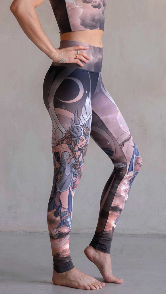Side view of model wearing WERKSHOP Valkyrie Athleisure Leggings. featuring original artwork by Chriztina Marie. featuring a Valkyrie warrior from Norse Mythology flying through a dark sky holding a shield and swords. Her hair is in braids and she is wearing armor and a viking helmet. The colors are warm and the background also features a crescent moon and stars.