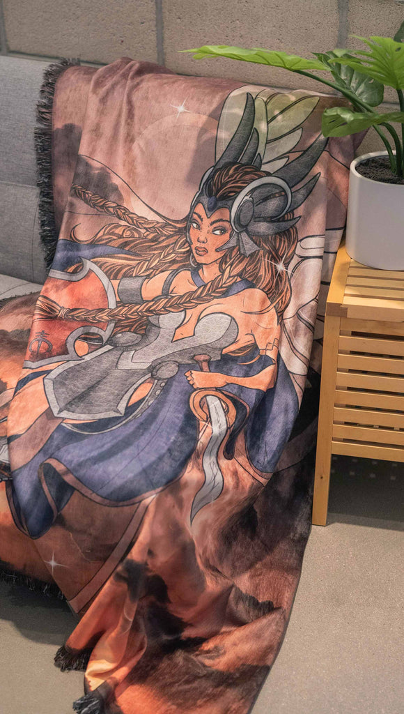 Decorative Chenille Tapestry printed with limited edition artwork by our Female Founder, Chriztina Marie. The artwork features a valyrkie warrior flying through a stormy sky holding a sword and shield. The colours are rich and warm with peach/orange tones and some pops of blue. This image shows the tapestry draped on a couch.