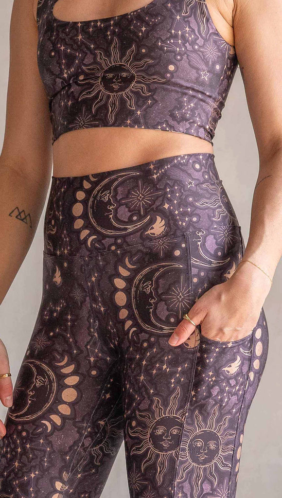 Model wearing WERKSHOP Zodiac Bell Bottoms. They are high waist and feature pockets on both legs. The zodiac themed artwork shows a hand-drawn sun and moon with the moon phases, shooting stars and all 12 zodiac constellations in gold over a dark purple background. Our model is 5’2” and this images shows her wearing the bells with sneakers. The hem of the pants are hitting the floor.