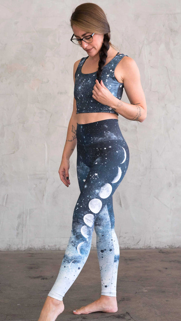 full body view of a girl wearing our moon phases athleisure leggings - with the phases of the moon printed on the wearer's left leg on a starry night sky with all zodiac constellations in the background
