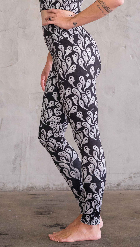 Enhanced left view of model wearing black athleisure leggings with little white ghosts and the word, "Boo!" in a repeat pattern