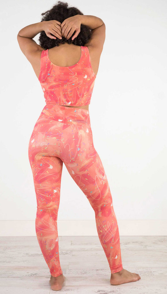 Full body back view of model wearing WERKSHOP Salmon Scribble Leggings with coral  brushstrokes over a bright salmon background. Also has little confetti and eagle logos scattered throughout.