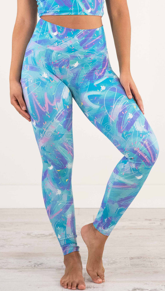 Front waist down view of model wearing WERKSHOP Teal Scribble Leggings with purple and pink brushstrokes over a bright teal background. Also has little confetti and eagle logos scattered throughout.