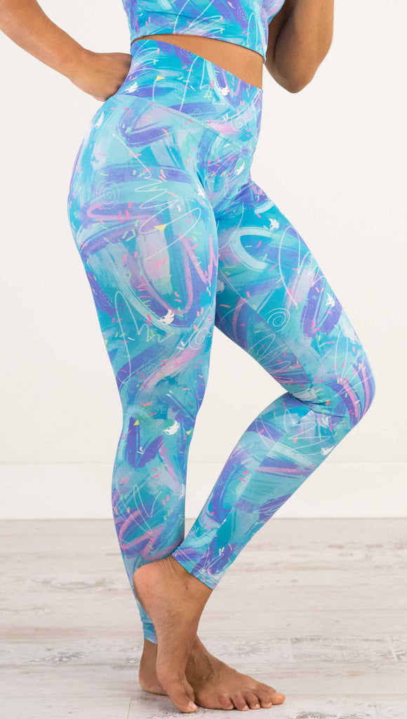 Waist down side view of model wearing WERKSHOP Teal Scribble Leggings with purple and pink brushstrokes over a bright teal background. Also has little confetti and eagle logos scattered throughout.