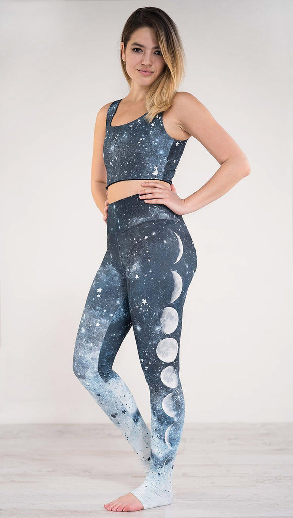 Left side view of model wearing blue full length athleisure leggings that fade to white with different moon phases in a vertical direction and tiny white stars across the leggings