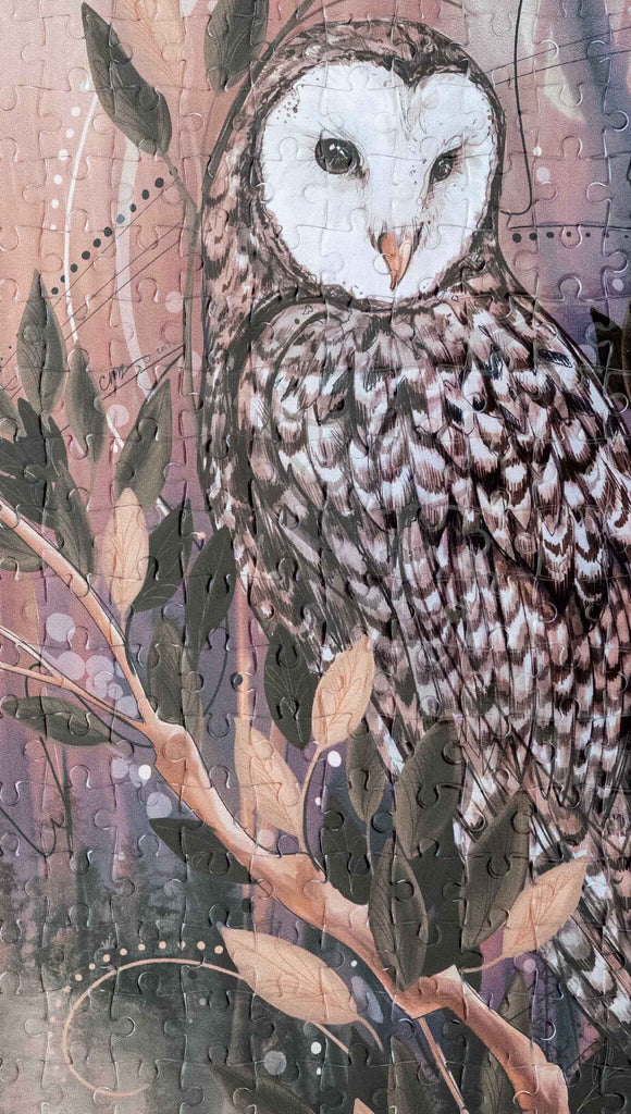 Zoomed in image of WERKSHOP Barn Owl 252 piece jigsaw puzzle. The puzzle is printed with original artwork of an Owl by Chriztina Marie. It features a barn owl perched on top of a branch with whimsical leaves. The overall color story is of mauves, warm browns and complimentary green leaves. 