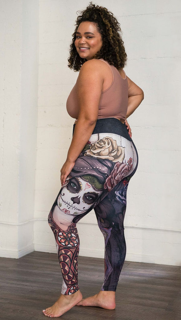 Three quarter rear right view of model wearing mashup Sugar Skull and Dia De Los Muertos themed printed full length leggings