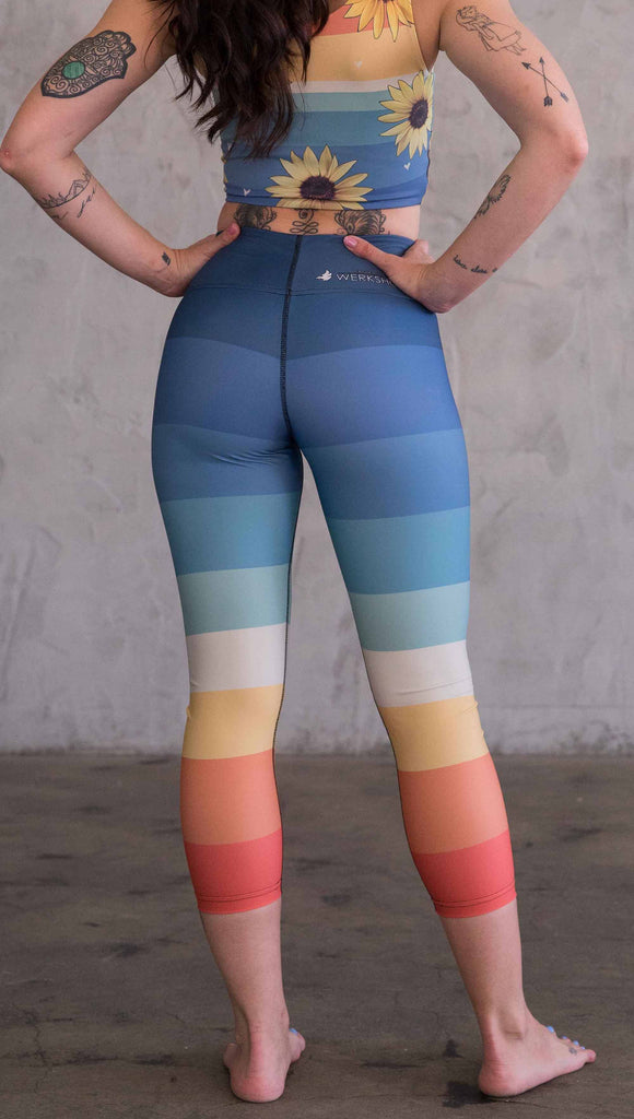 full body back view of model wearing WERKSHOP Vintage Rainbow Triathlon Capri leggings. The leggings have wide horizontal stripes with dark blue at the waistband, to auqua and pale green at the mid thigh leading to cream at the knee and orange and red tones to the ankle.
