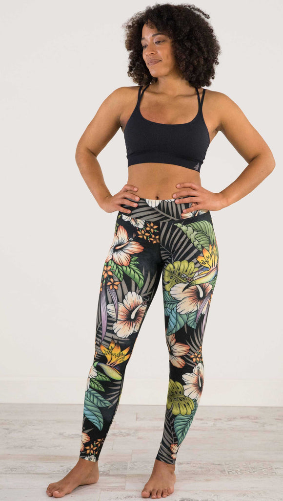 Full body front view of model wearing WERKSHOP Floral Night Full Length Triathlon Leggings. The artwork on the leggings has tropical flowers (bird of paradise, hibiscus and palm leaves) on a distressed black background.