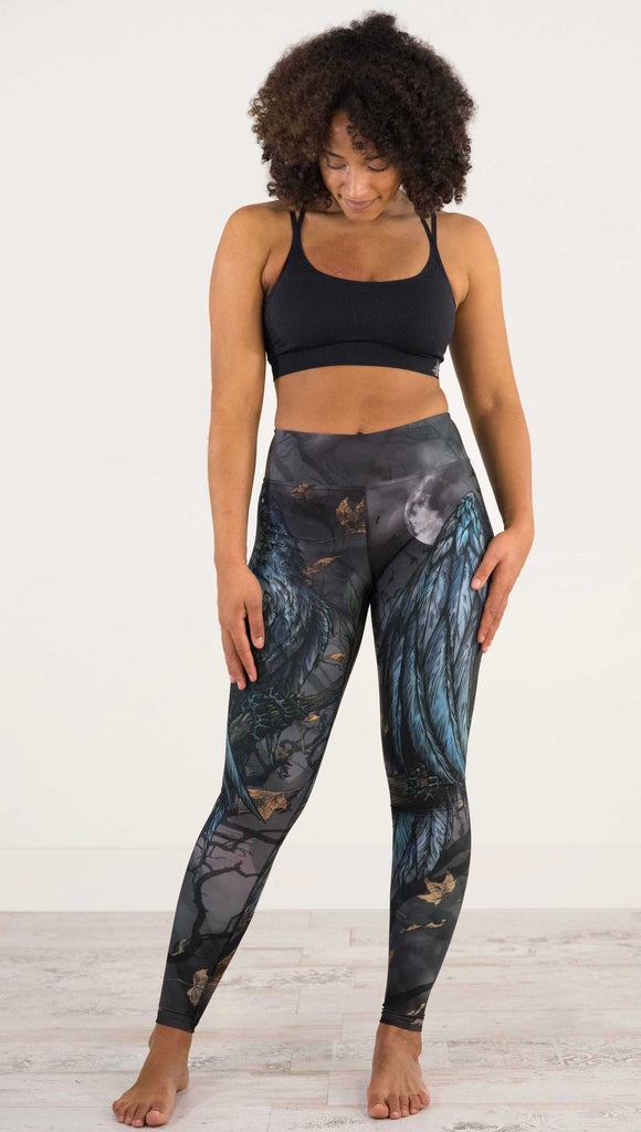 Full body front view of model wearing WERKSHOP Raven Full Length Triathlon Leggings. The artwork on the leggings is super dark shades of gray and blue with a large raven on the thigh and a dark and stormy moon in the background. Definitely spooky season vibes.