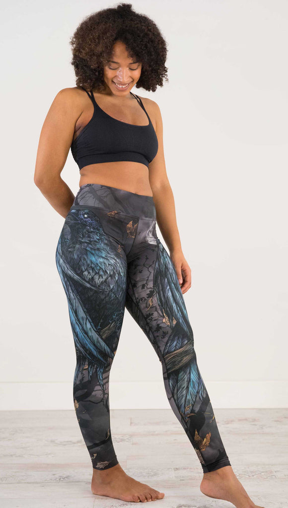 Full body side view of model wearing WERKSHOP Raven Full Length Triathlon Leggings. The artwork on the leggings is super dark shades of gray and blue with a large raven on the thigh and a dark and stormy moon in the background. Definitely spooky season vibes.