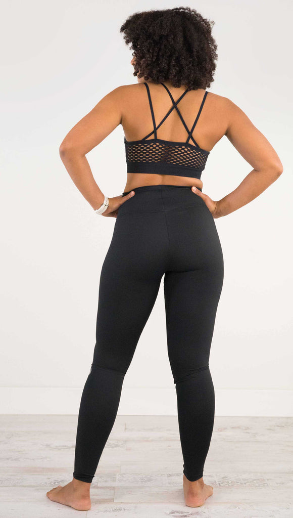 Full body back view of model wearing WERKSHOP Solid Black Athleisure Leggings with a small reflective eagle logo on the wearers left side calf