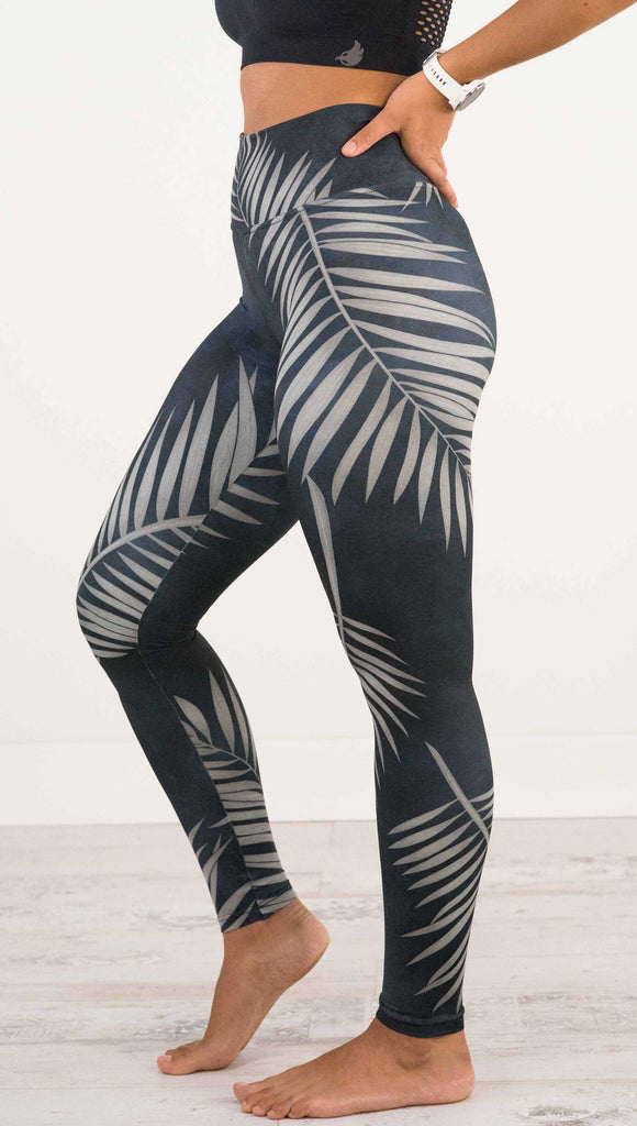Waist down side view of model wearing WERKSHOP Black Palms leggings. The leggings are printed with a mostly black watercolor effect with grayish palm frond silhouette wrapping the body.