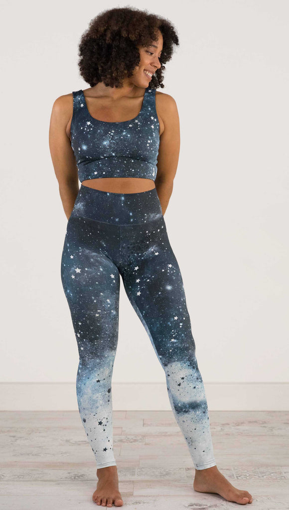 Full body front view of a girl wearing our moon phases athleisure leggings - with the phases of the moon printed on the wearer's left leg on a starry night sky with all zodiac constellations in the background