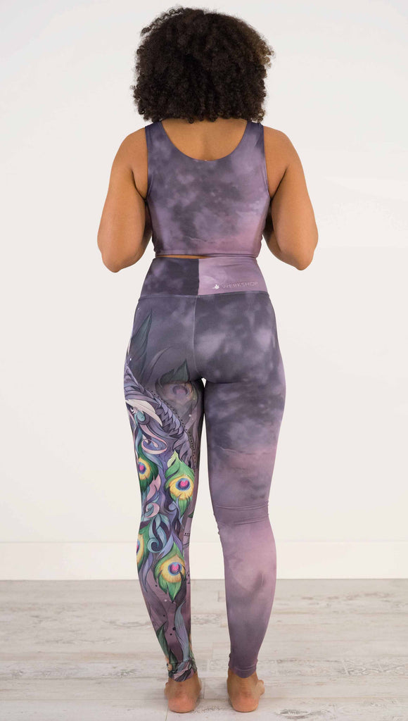 Full body back view of model wearing WERKSHOP Peacock Athleisure Leggings. The artwork on the leggings features a vibrant, colorful peacock down the wearer's left leg over a beautiful warm purple and pink watercolor background.