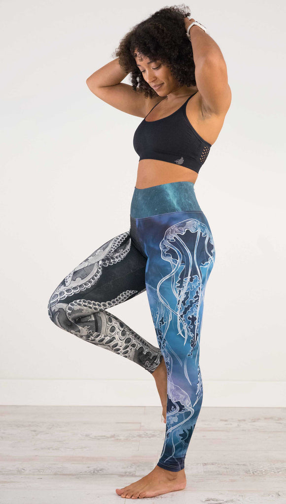 Full body side view of model wearing WERKSHOP Sea Mashup Athleisure Leggings. The artwork on the leggings features hand-drawn black and white tentacles wrapping up the wearers right leg and jellyfish over a blue watercolor background on the left leg. They are tied together with a blue-green under-the-sea themed waistband,