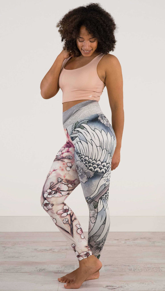 Full body side view of model wearing WERKSHOP Zen Mashup Athleisure Leggings. The leggigns are printed with pink cherry blossoms on the wearer's left leg and a graceful swooping crane on the opposite leg. There is also an abstract outline drawing of a koi fish on the waistband.