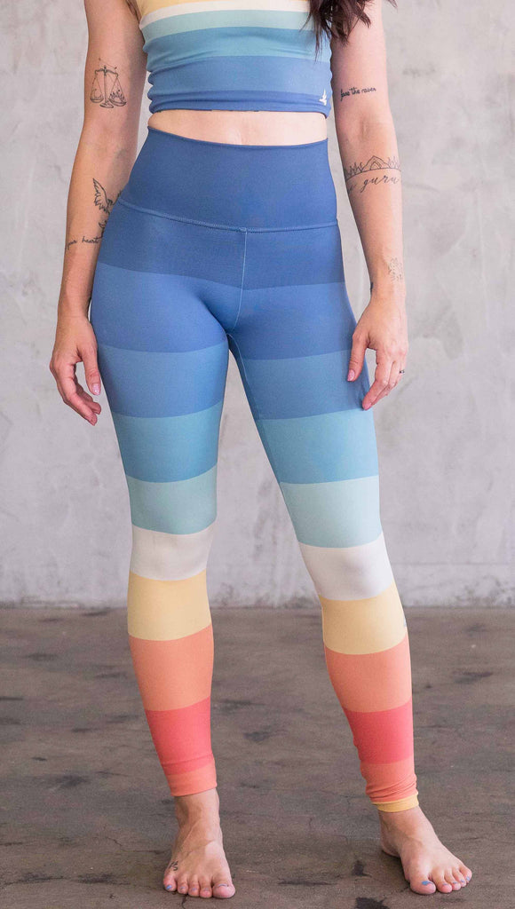 Waist down front view of model wearing WERKSHOP Vintage Rainbow Athleisure leggings. The leggings have wide horizontal stripes with dark blue at the waistband, to auqua and pale green at the mid thigh leading to cream at the knee and orange and red tones to the ankle.