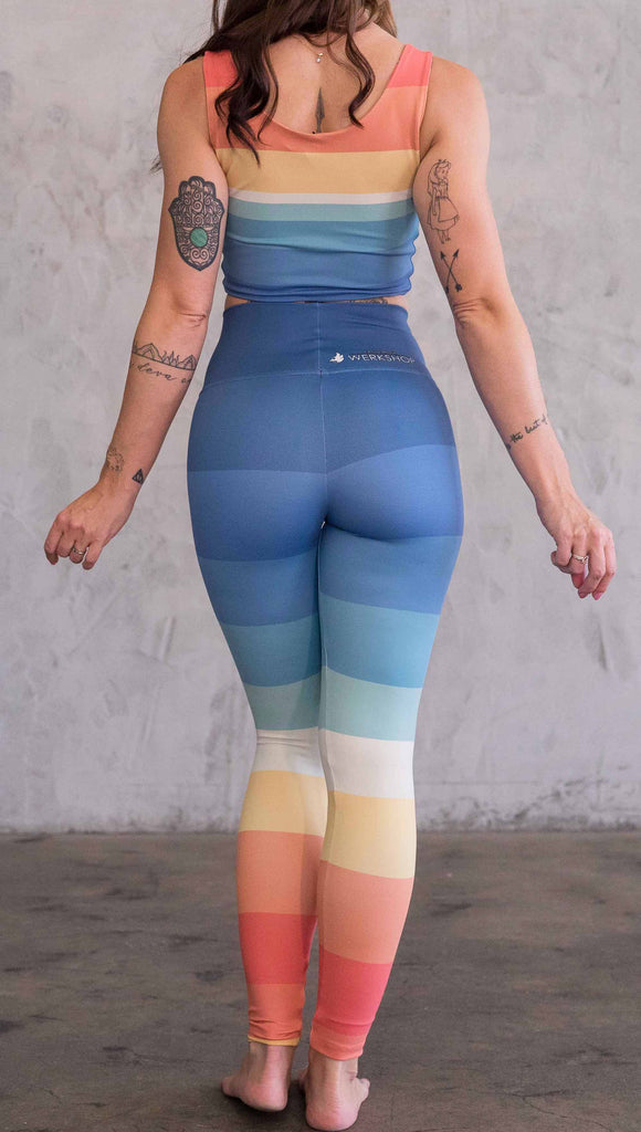 Full body back view of model wearing WERKSHOP Vintage Rainbow Athleisure leggings. The leggings have wide horizontal stripes with dark blue at the waistband, to auqua and pale green at the mid thigh leading to cream at the knee and orange and red tones to the ankle.