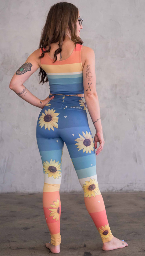 Full body back view of model wearing WERKSHOP Sunflower Athleisure leggings. The leggings have wide horizontal stripes with dark blue at the waistband, to aqua and pale green at the mid thigh leading to cream at the knee and orange and red tones to the ankle. There are large photo-real sunflowers and tiny hand sketched off-white hearts sprinkled throughout.