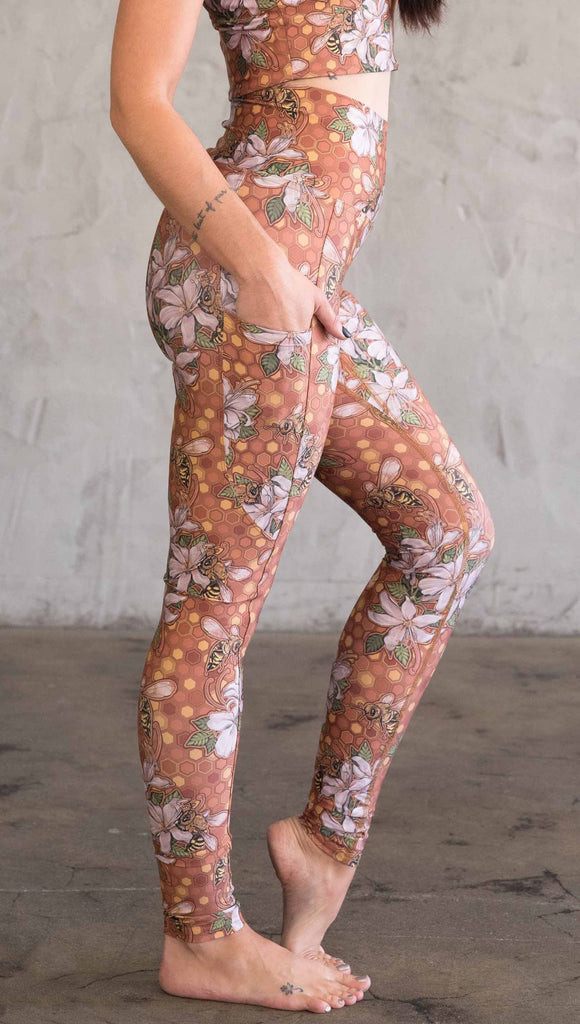 Side view of model wearing ultra lightweight "featherlight" leggings with clusters of honeybees and flowers. Her hand is in the side pocket that is big enough to hold a phone. 